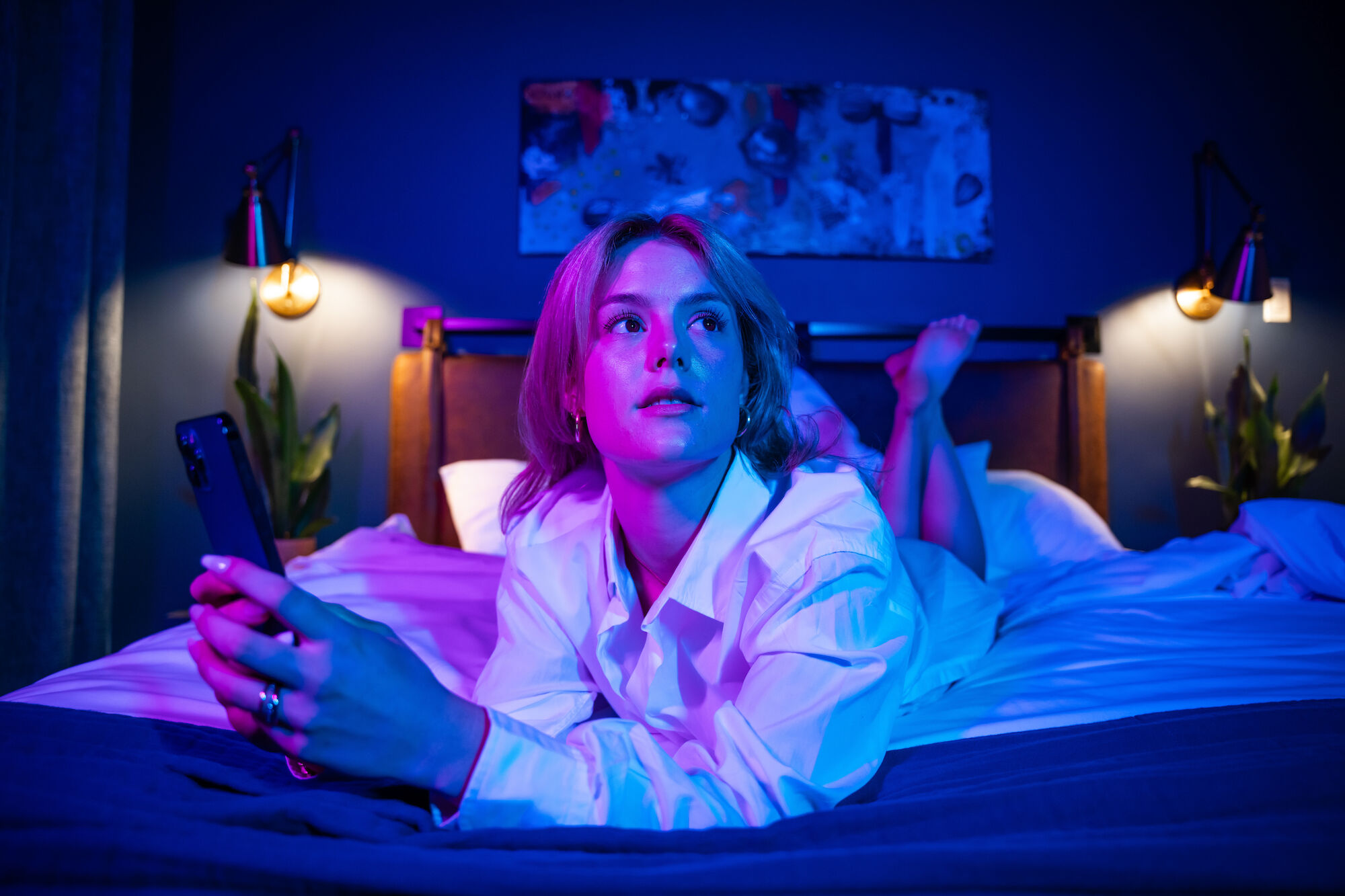 A person is lying on a bed, holding a phone, illuminated by purple and blue lighting, with wall art and lamps in the background.