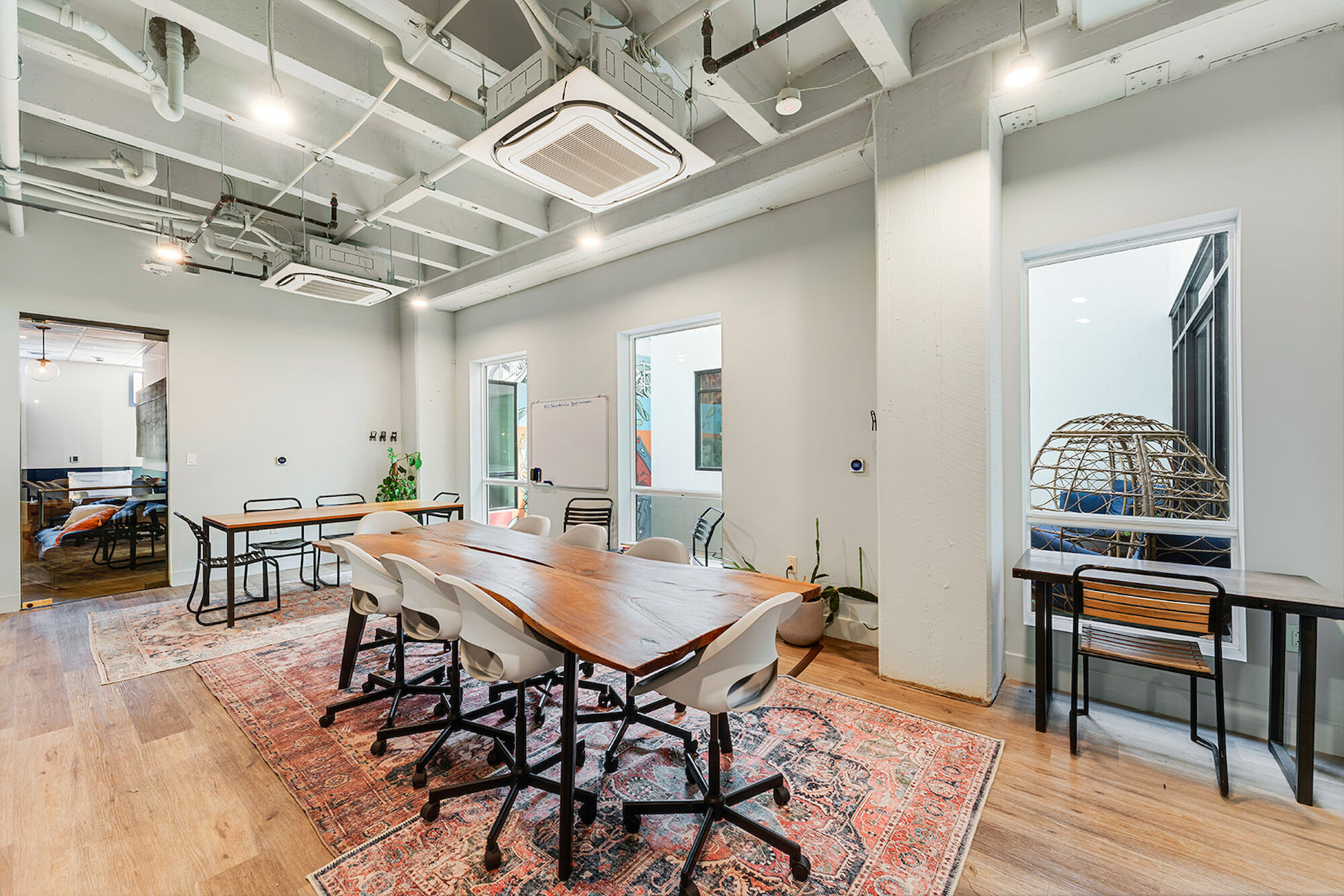 A modern conference room with a large wooden table, chairs, a rug, and an adjacent windowed room with a unique chair. The room is well-lit.