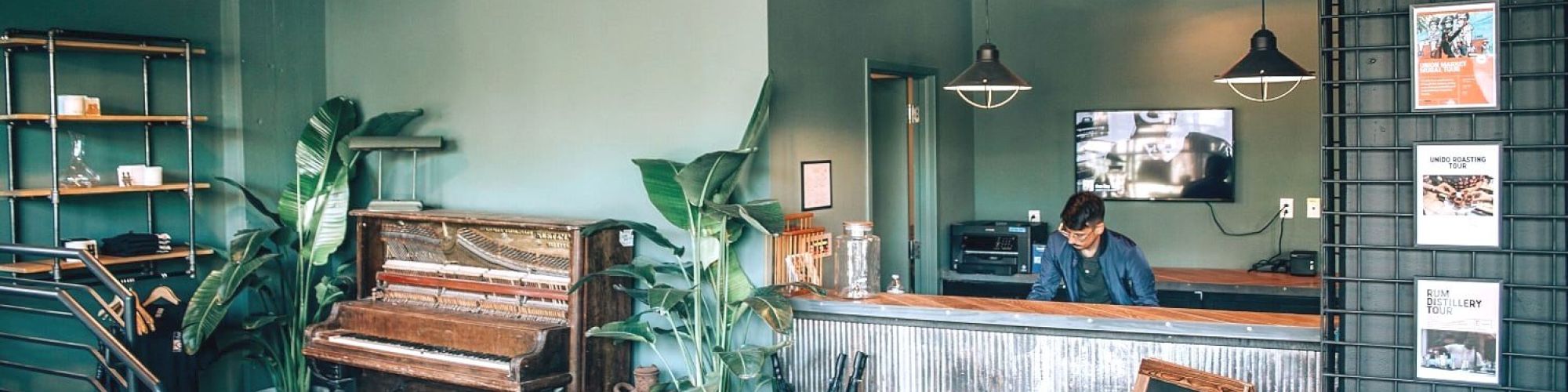 A modern reception area with a piano, plants, and decor. A person is behind the front desk, with a chalkboard sign and industrial lighting.