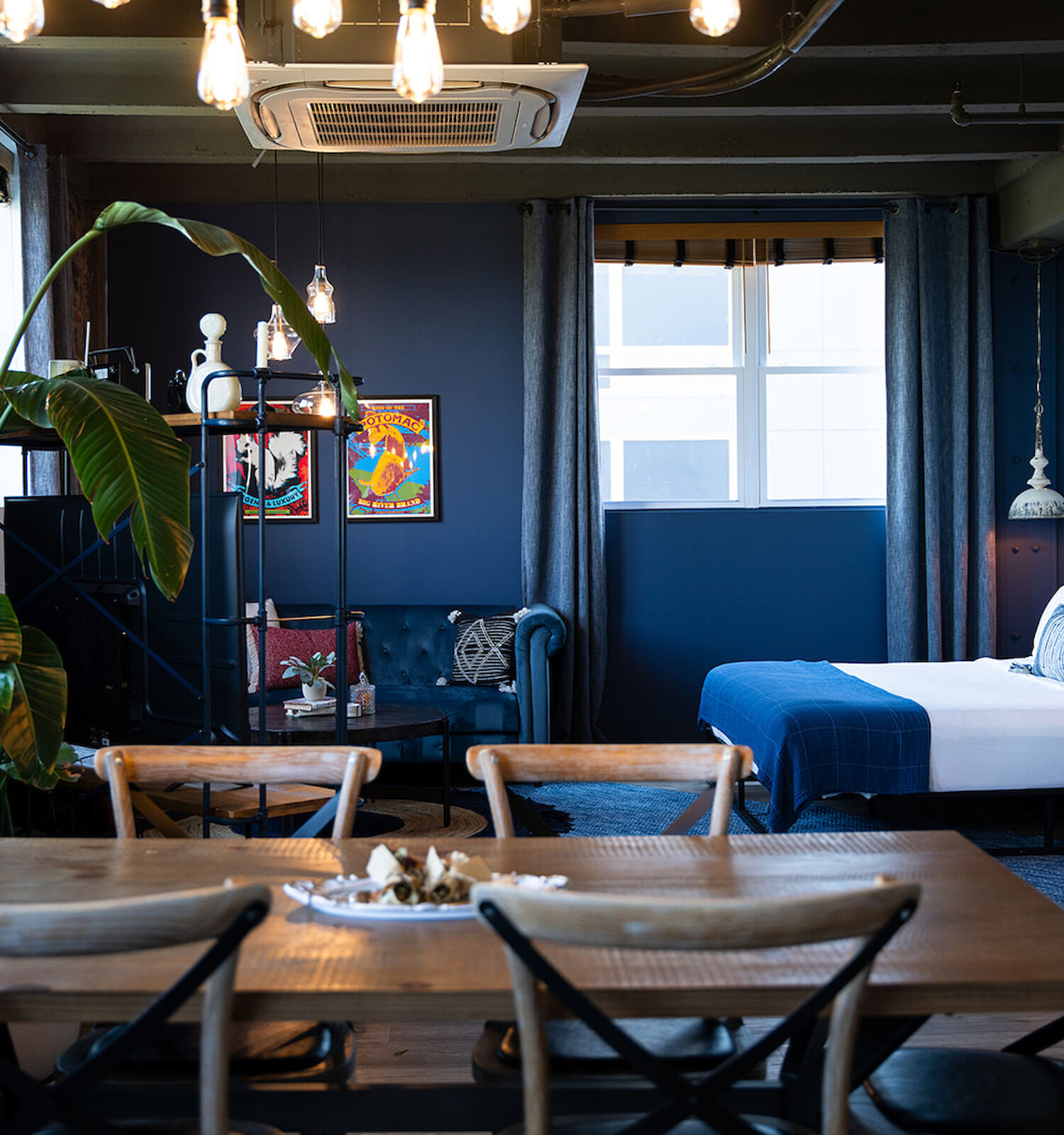 A modern room with a bed, dining area, large plant, wall art, and hanging lights, featuring a mix of wooden and industrial decor elements.