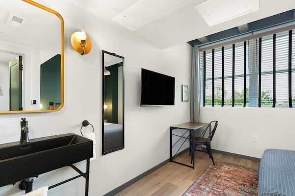 The image shows a small, modern room with a black sink, a large mirror, a wall-mounted TV, a desk with a chair, and a window with blinds.