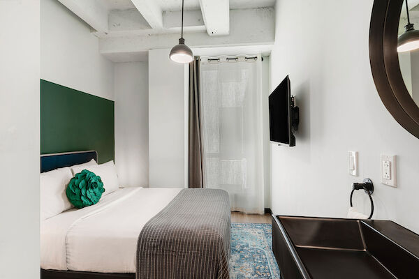 A modern bedroom with a neatly made bed, wall-mounted TV, round mirror, pendant light, and a sink. The room is decorated in a minimalist style.