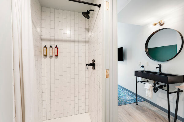 A modern bathroom with a walk-in shower, subway tiles, black fixtures, a black sink vanity, round mirror, and toiletries on the wall.