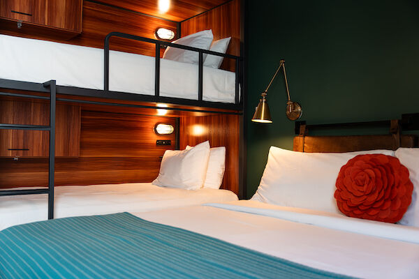 The image shows a cozy room with a bunk bed and a double bed, featuring white linens, a red decorative pillow, a blue throw blanket, and warm lighting.