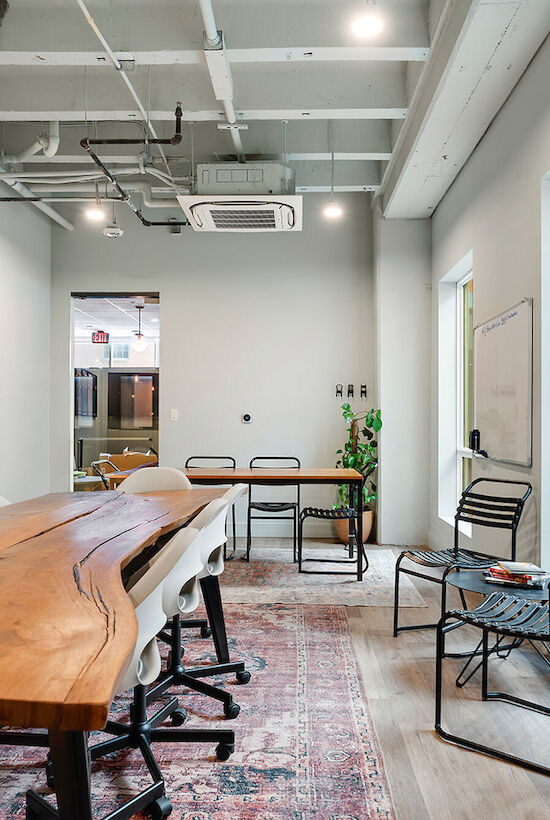 A modern conference room with a large wooden table, several chairs, wall art, a potted plant, a whiteboard, and ample natural light from windows.