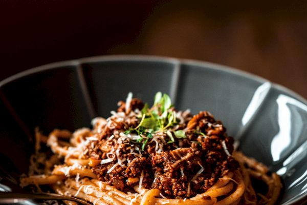 A hand holding a black bowl of spaghetti topped with a meat sauce and green garnish, set against a dark, out-of-focus background.