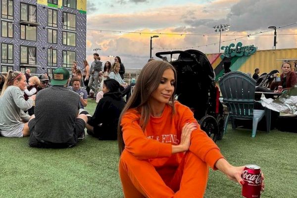 A woman in an orange tracksuit sits on the grass, holding a can of Coca-Cola, with a group of people and a tall building in the background.