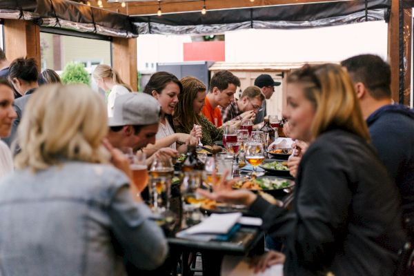 A group of people are gathered around a table, enjoying food and drinks at an outdoor setting, suggesting a casual gathering or social event.