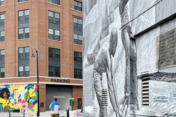 A person and a dog walk on a path beside a street art mural of dancers on a building wall. In the background, there's a colorful mural and modern buildings.