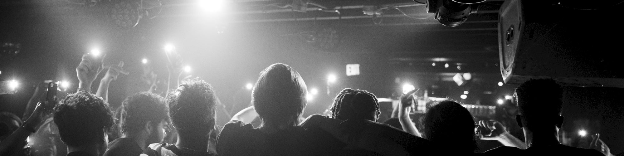 A group of people with their backs to the camera embrace under bright stage lights, holding up their phones to capture the moment.