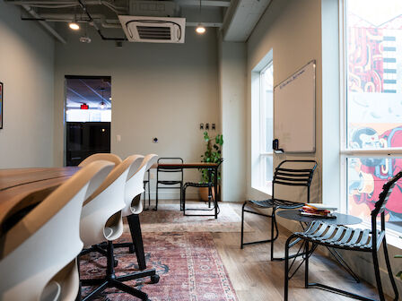 A modern conference room with a long table, office chairs, wall art, a whiteboard, additional seating, and large windows displaying an outside mural.