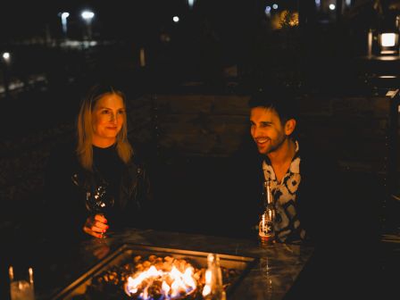 Two people are sitting outdoors by a fire pit at night, enjoying drinks, and having a conversation.