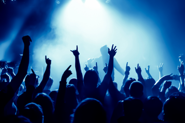 A crowd at a concert, cheering with hands raised, illuminated by vibrant blue stage lights and a haze of smoke.