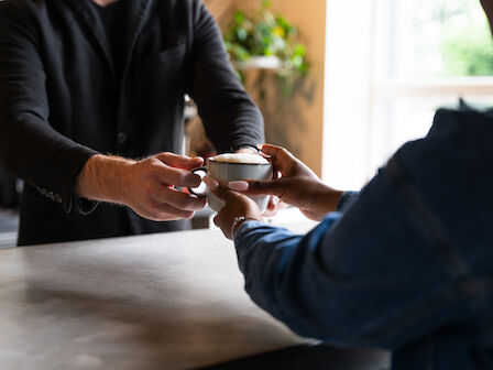 Two people exchanging a cup of coffee across a counter in a brightly lit room with plants in the background.