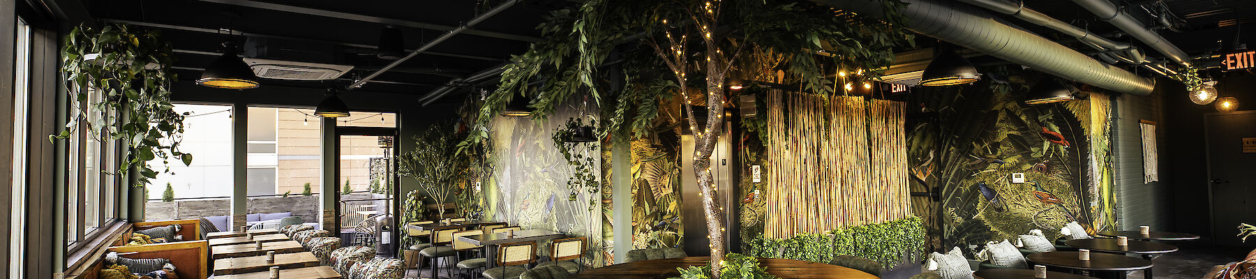 The image shows a modern restaurant interior with green decor, plants, various seating options, and warm lighting, creating a cozy atmosphere.