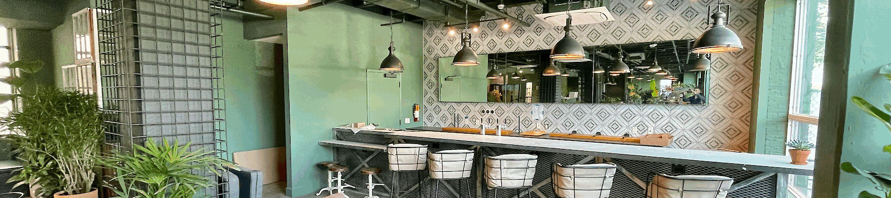 The image shows a modern, stylish indoor space with seating, a bar area with stools, and green plants. There are pendant lights hanging from the ceiling.