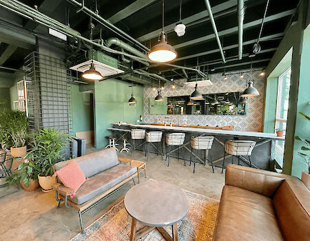 The image shows a modern, stylish indoor space with seating, a bar area with stools, and green plants. There are pendant lights hanging from the ceiling.