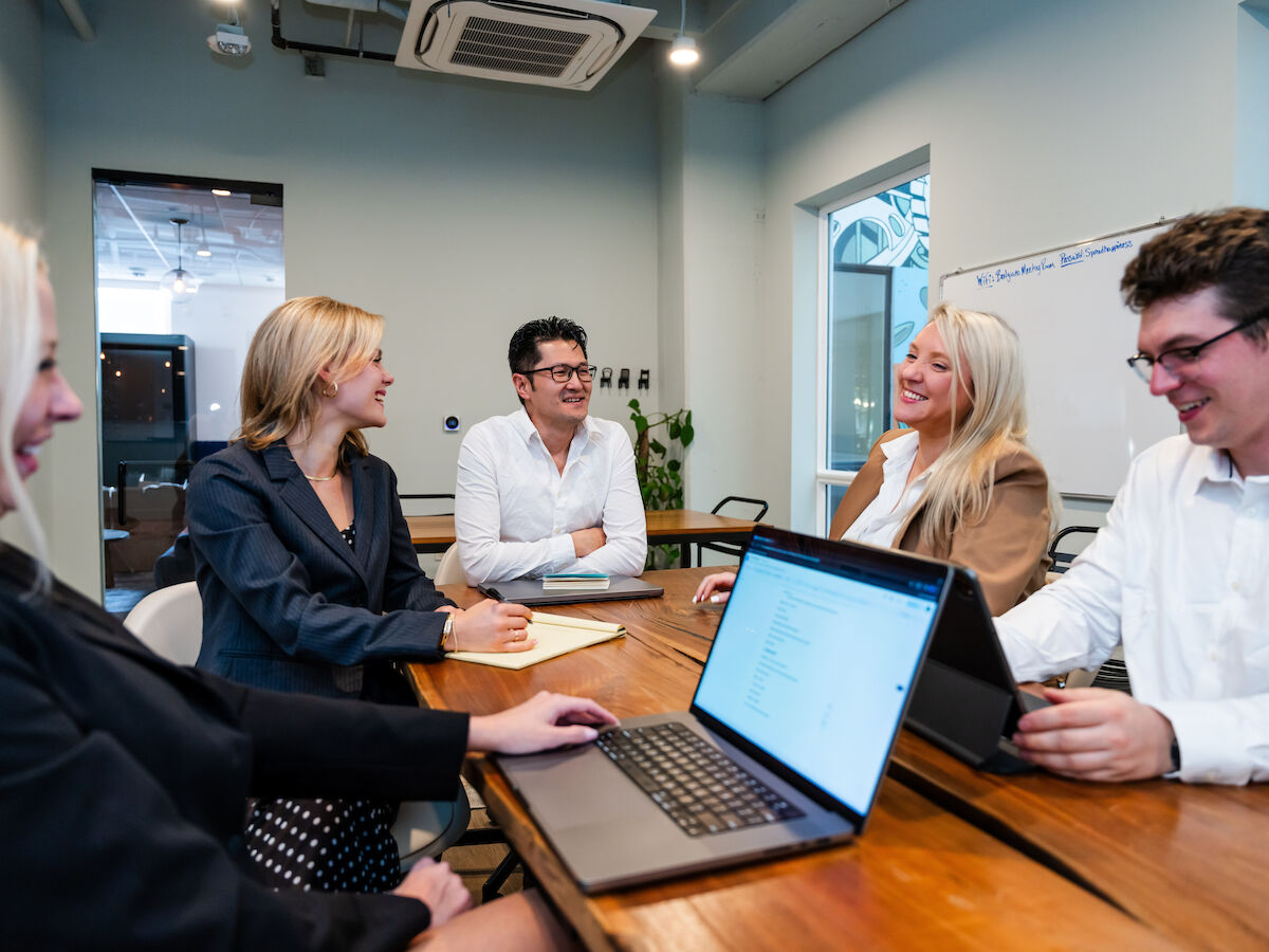 A group of five people is having a meeting in a conference room, with laptops and notebooks on the table, and they appear to be engaged in discussion.