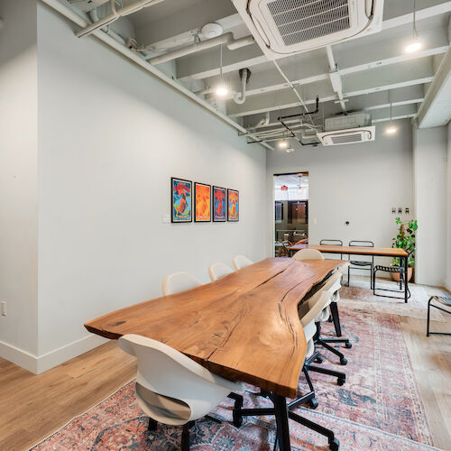 A modern conference room with a wooden table, chairs, wall art, bookshelves, and indoor plants, featuring a light industrial ceiling design.