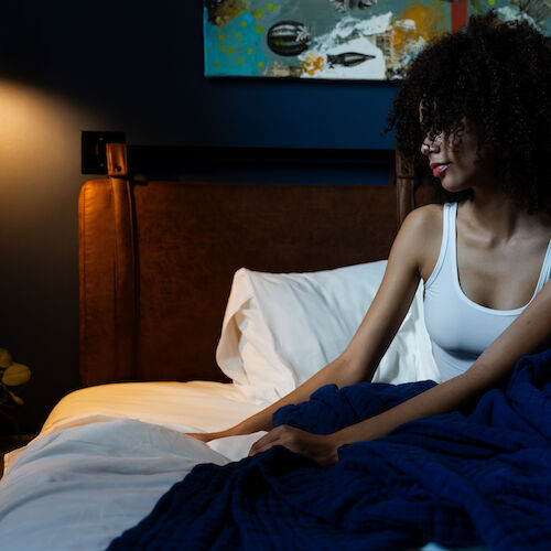 A person sits on a bed, partially covered by a blue blanket in a dimly lit room, with a bedside table, wall lamp, and artwork in the background.