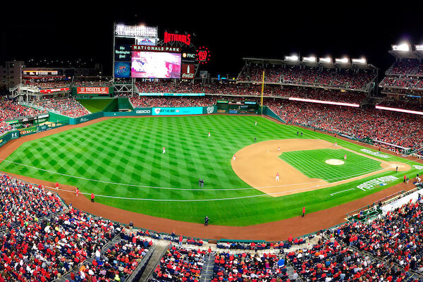 A wide-angle view of a brightly lit baseball stadium at night, filled with a large crowd, showcasing the field and the scoreboard in the background.