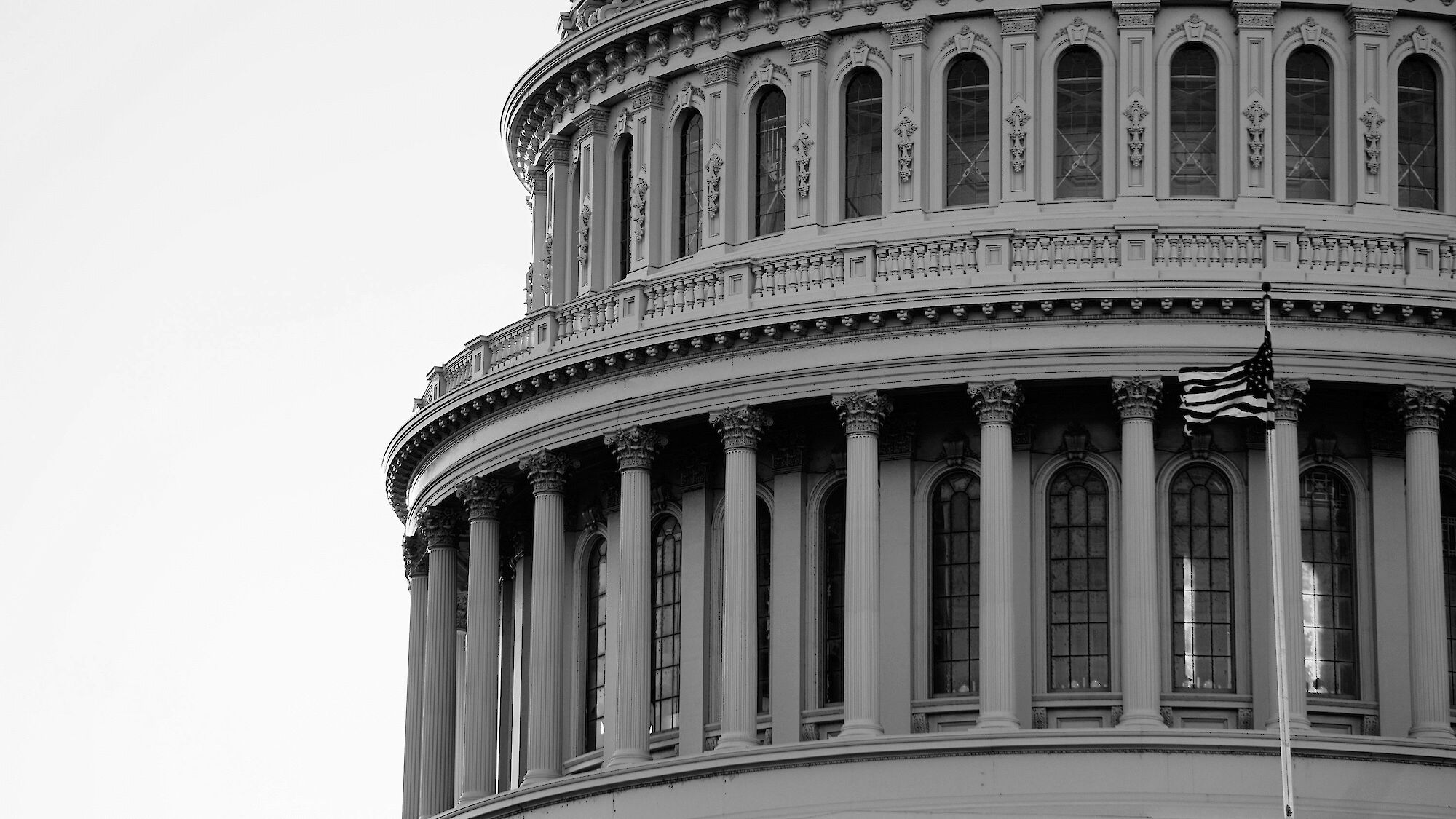 This black-and-white image shows a close-up view of the dome of the U.S. Capitol building, featuring columns and a partially visible American flag.