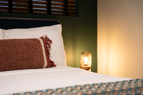 A cozy bedroom with a bed, a patterned throw pillow, a bedside lamp, white bedsheets, and a window with wooden shutters in the background.