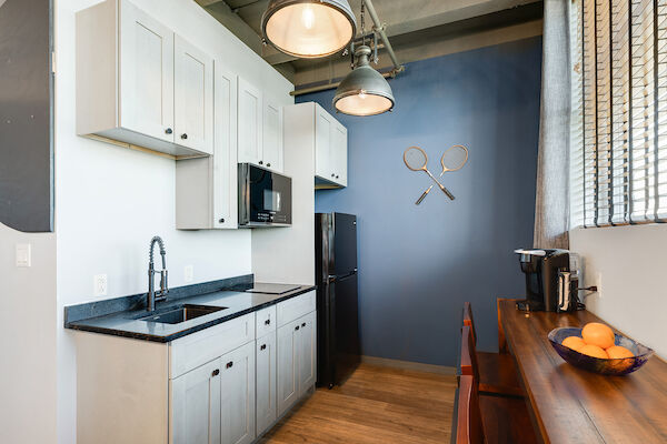 A modern kitchen with white cabinets, black countertops, a black fridge, and a microwave, features wall decor of two tennis rackets crossed.