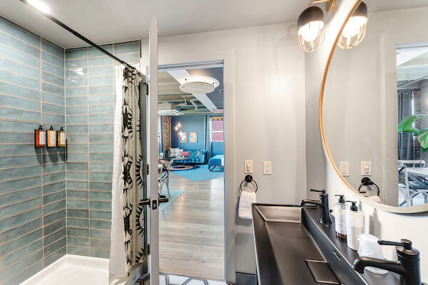 The image shows a modern bathroom with a shower, a vanity with two sinks, and a round mirror, opening to a living area.