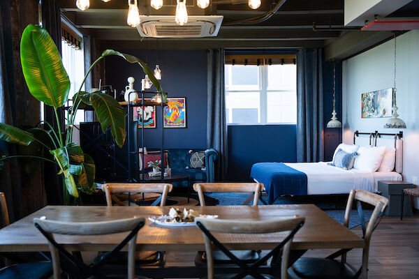 This image shows a modern, stylish room with a bed, dining area, plants, wall art, and natural light from windows.