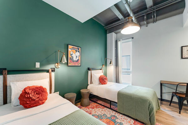 The image shows a well-lit room with two single beds, green accent walls, red pillows, a small desk, and modern décor, creating a cozy ambiance.