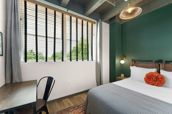 A bedroom with green walls, a desk and chair, bed with gray bedding and a red pillow, large window with blinds, and a ceiling light fixture.