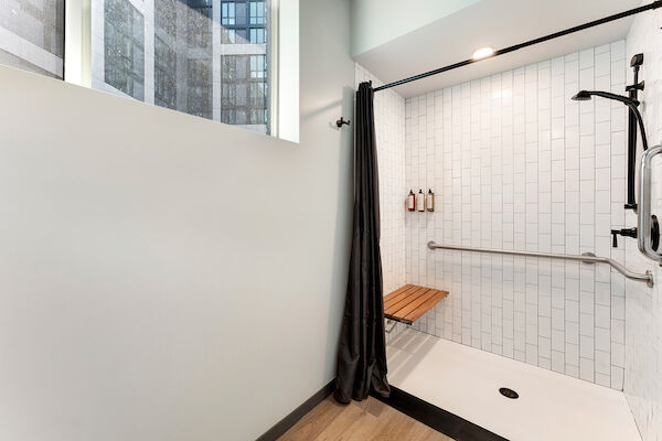 A modern, accessible shower with a bench, grab bar, and adjustable showerhead, featuring white tile walls and a black curtain.