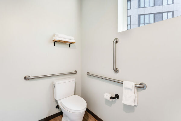 A clean, modern bathroom with a toilet, grab bars, a towel rack with towels, and a window with a view of a building outside.