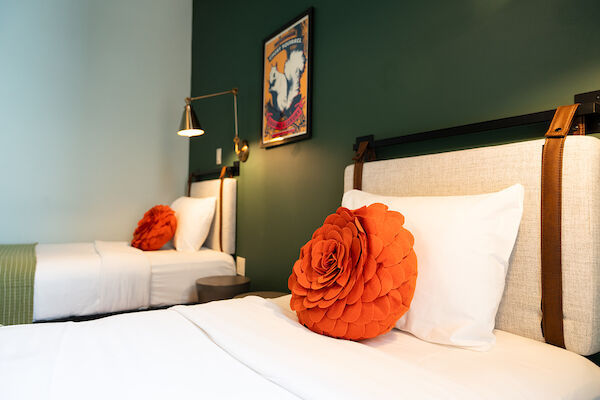 Two beds with white linens and large orange flower pillows, green-painted wall, framed artwork, and a wall lamp in a modern bedroom.