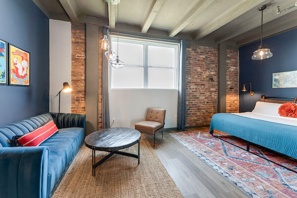 The image shows a modern bedroom with a blue sofa, a round coffee table, a wooden chair, wall art, and a neatly made bed with blue bedding.