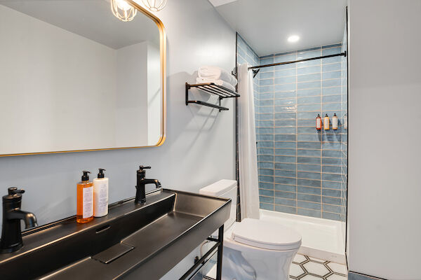 The image shows a modern bathroom with a double sink vanity, a large mirror, a toilet, and a shower with blue tiles.