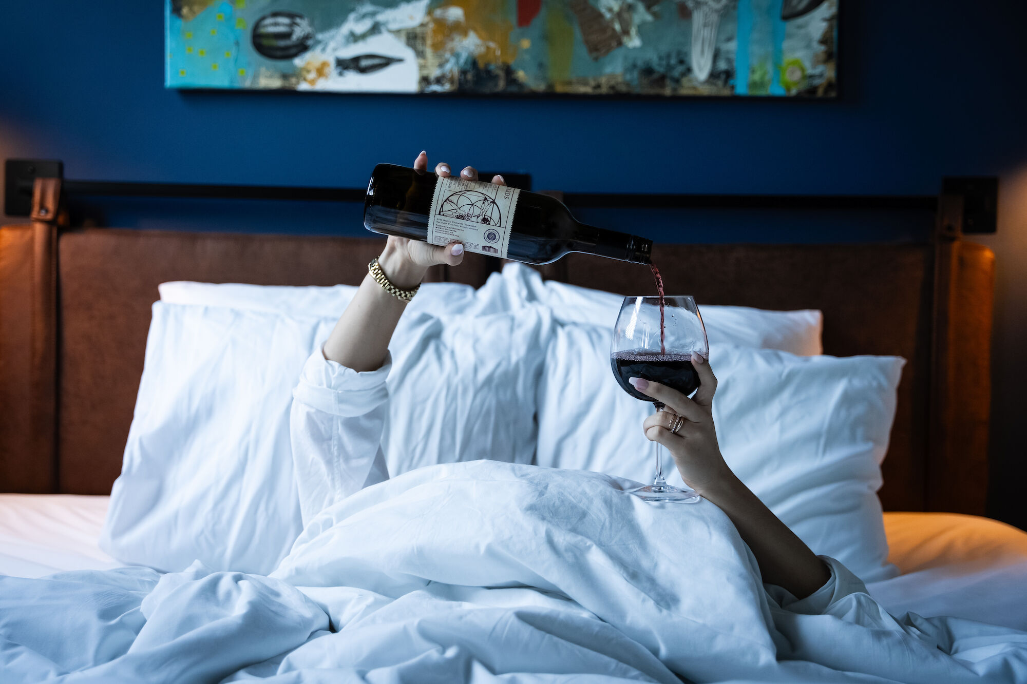 A person in white sleeves is lying in bed, pouring red wine from a bottle into a glass. The scene is relaxing and cozy.