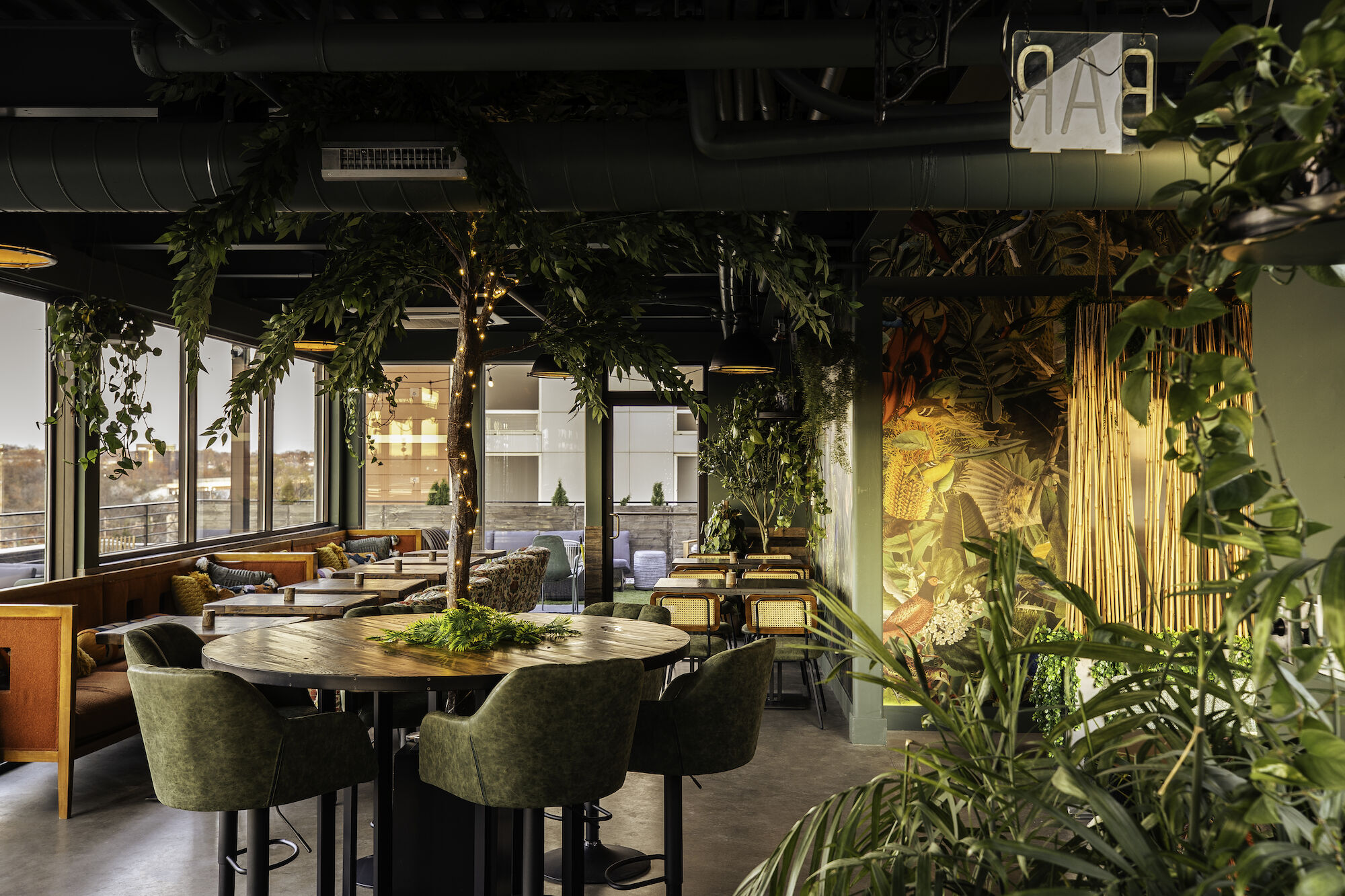 A beautifully designed restaurant interior with abundant greenery, modern furniture, large windows, and ambient lighting, creating a cozy atmosphere.