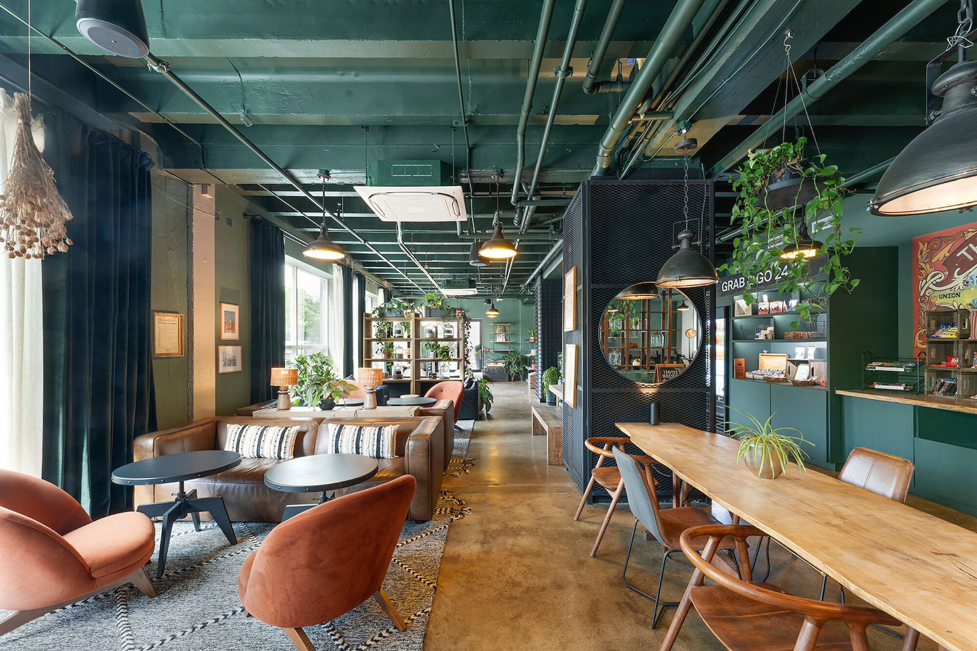 This is an image of a stylish, modern coffee shop with various seating options, green walls, hanging plants, and industrial lighting.