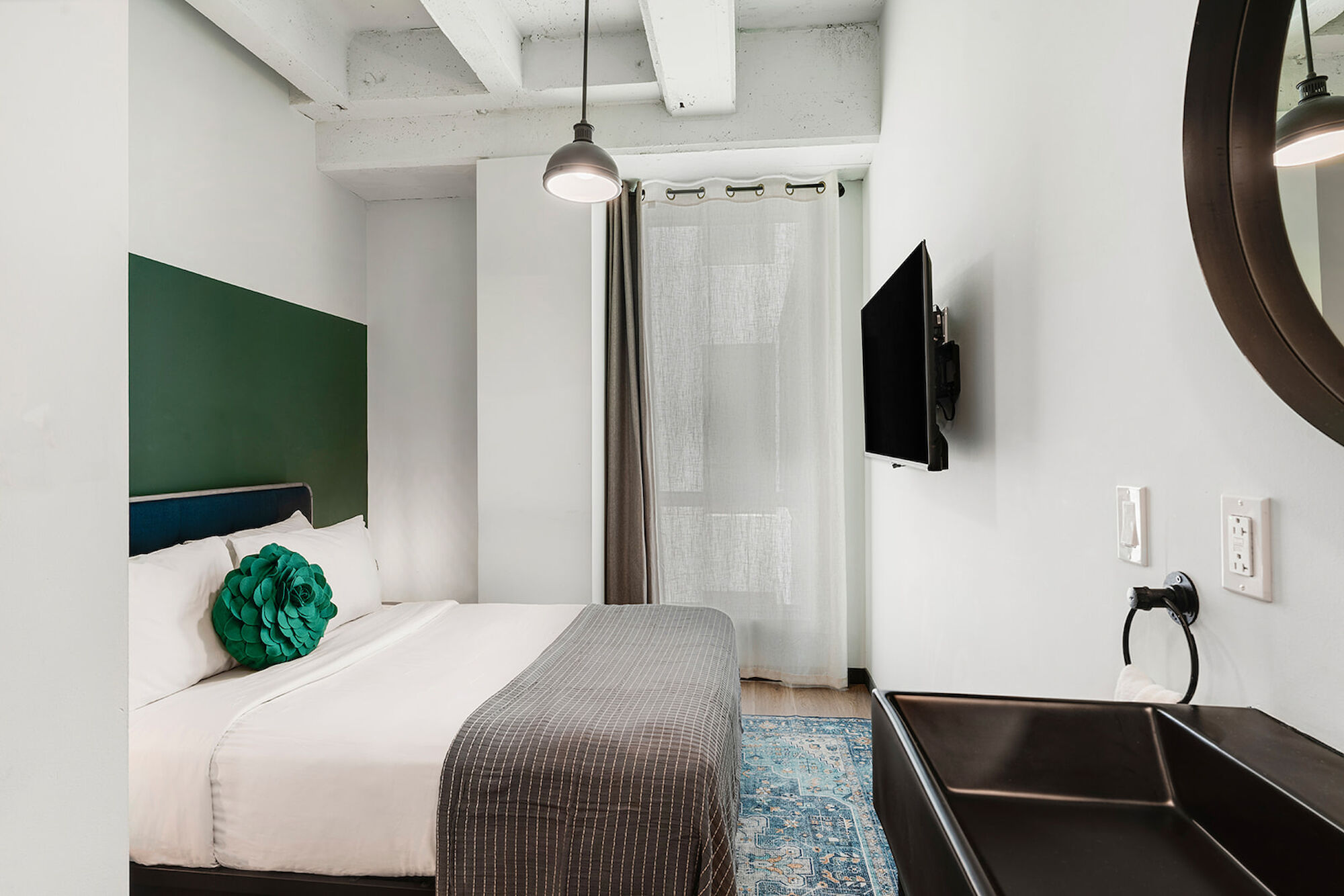 A modern bedroom with a bed, pillows, a TV mounted on the wall, a mirror, a rug, and curtains. The room has a minimalist design.