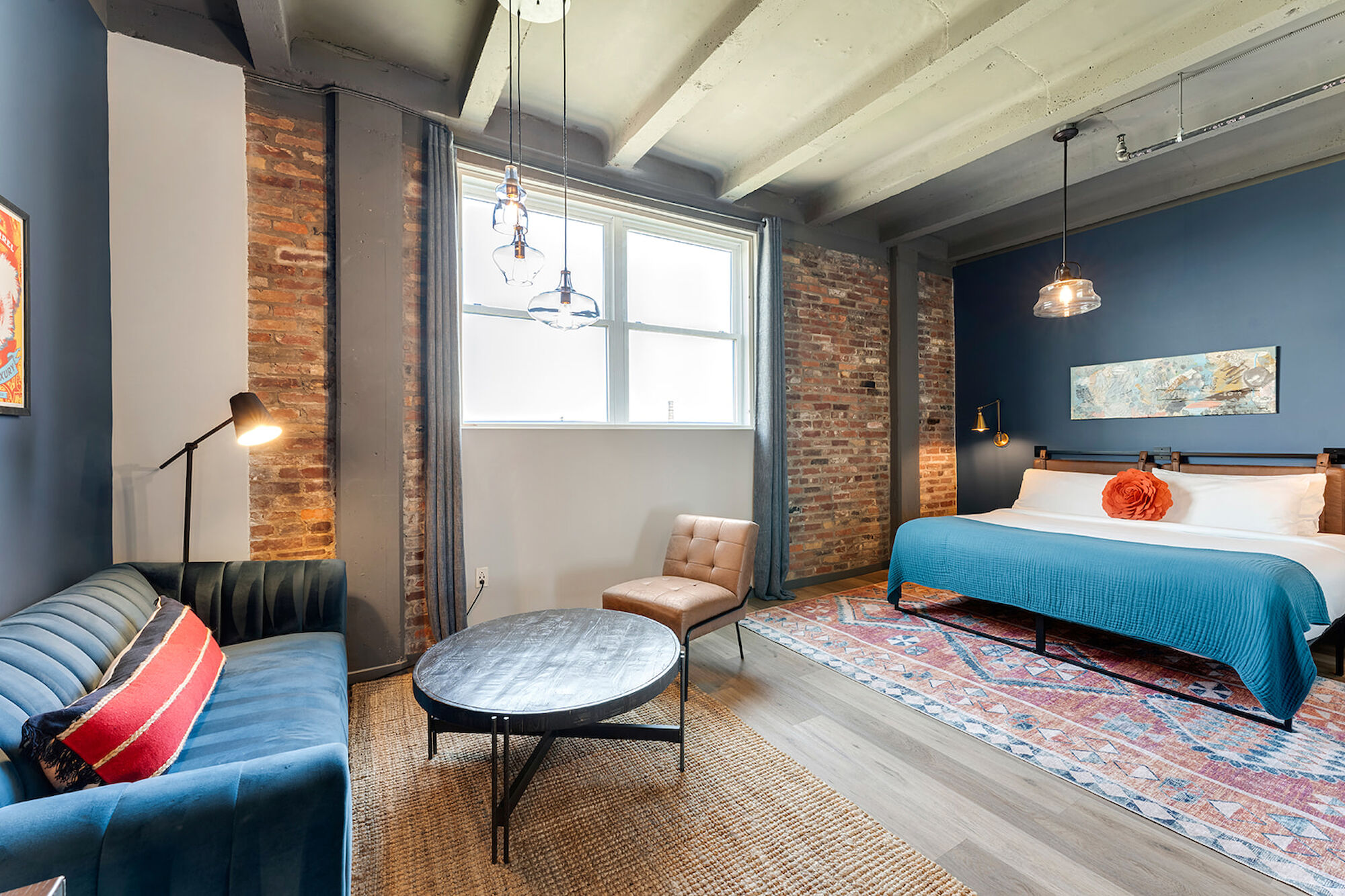 A modern room with a bed, couch, chair, coffee table, and stylish decor, featuring exposed brick walls and a large window.