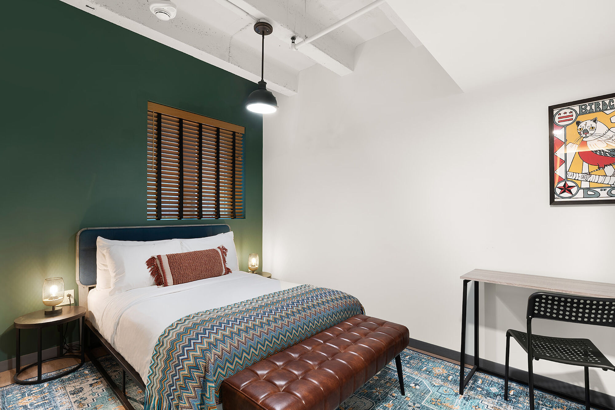 A cozy bedroom with a green accent wall, bed, bench, artwork, desk, and chair. Two bedside lamps are also visible in the room.