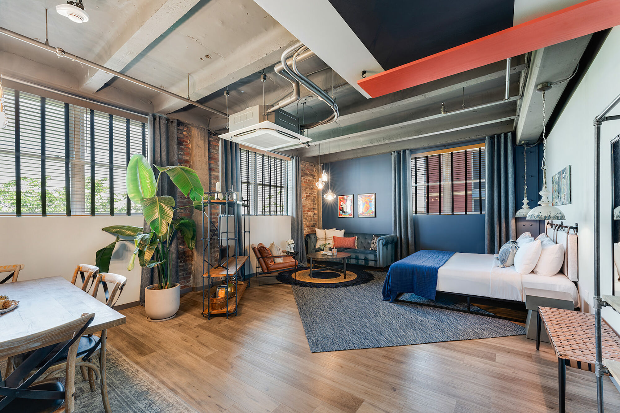 A modern loft with a mix of wood and metal elements, featuring a bed, couch, plants, and a small dining area under large windows, ending the sentence.