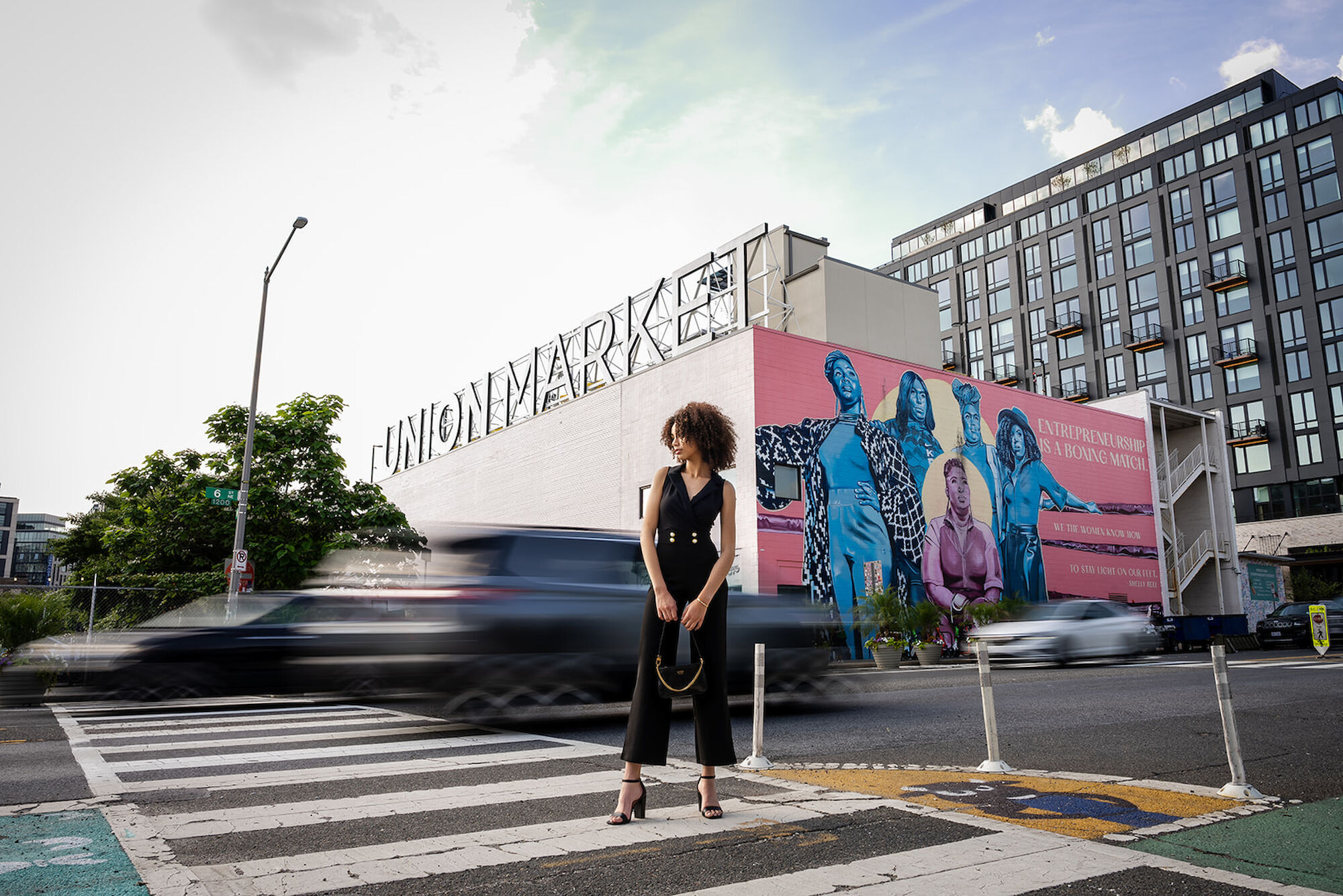 A woman stands at a crosswalk in front of a building with a large mural and a sign that reads 