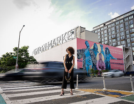 A person stands on a crosswalk near Union Market with a colorful mural and buildings in the background, as cars pass by.