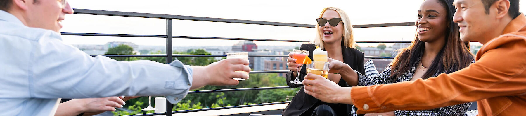 A group of four people are sitting outside, toasting drinks and enjoying food on a table with a scenic view in the background.