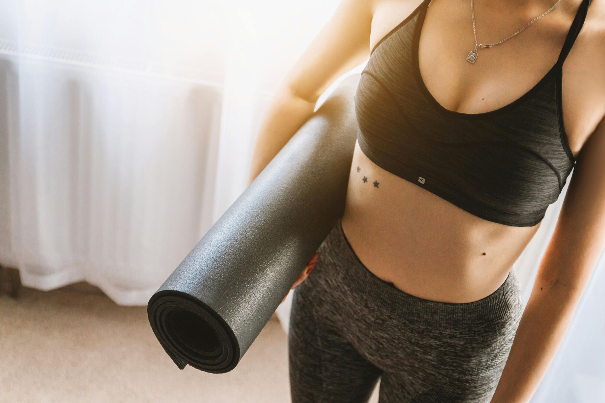 A person in athletic wear holds a rolled-up yoga mat, standing indoors with sunlight streaming in through the window.