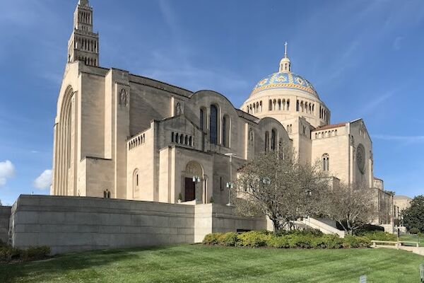 The image shows a large cathedral with a prominent dome and tower, set against a clear blue sky, surrounded by green lawns and trees.